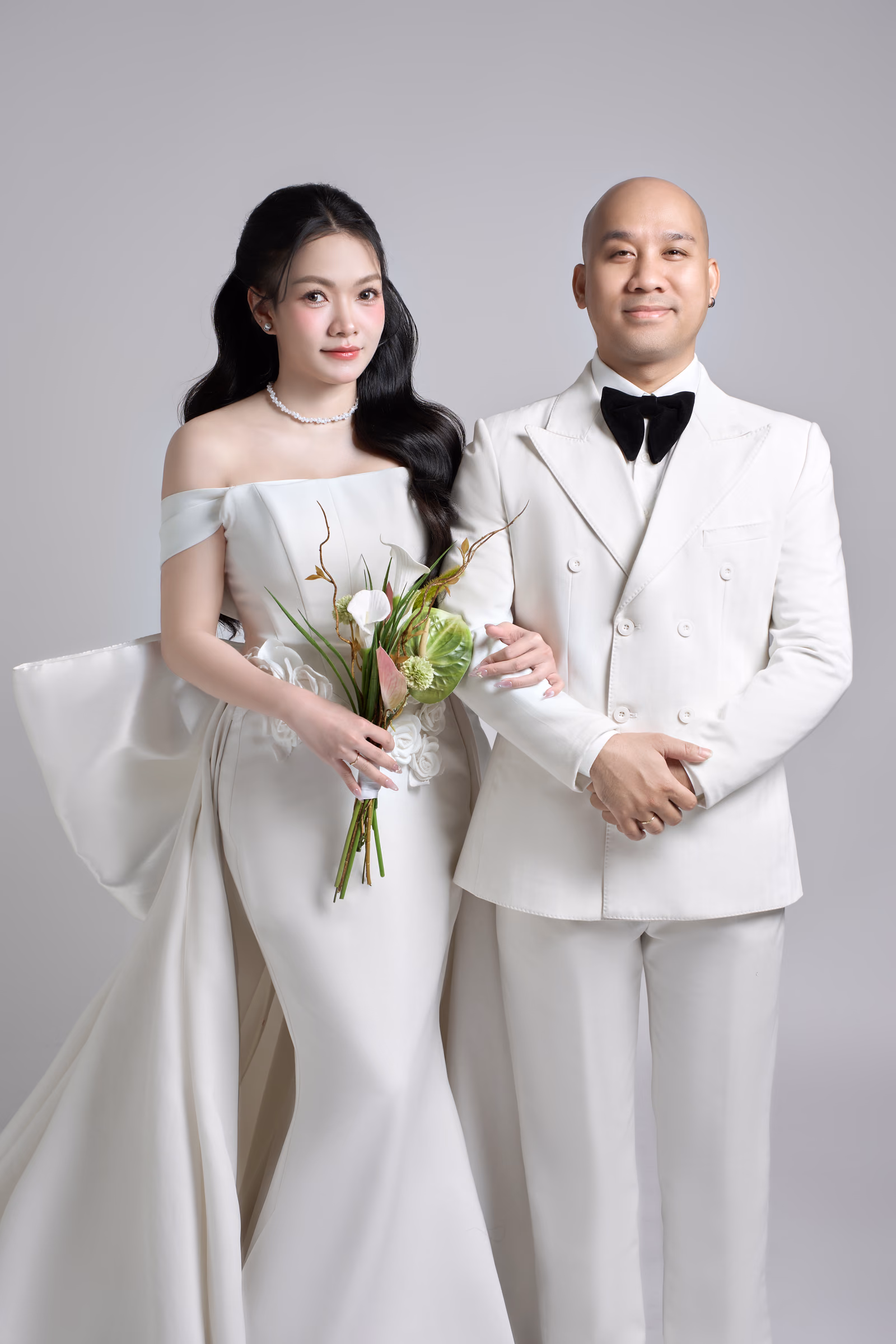 Cô dâu váy cưới trắng trễ vai cầm bó hoa calla nhỏ đứng cạnh chú rể mặc vest trắng thắt nơ đen, cả hai nhìn thẳng trên nền xám nhạt.