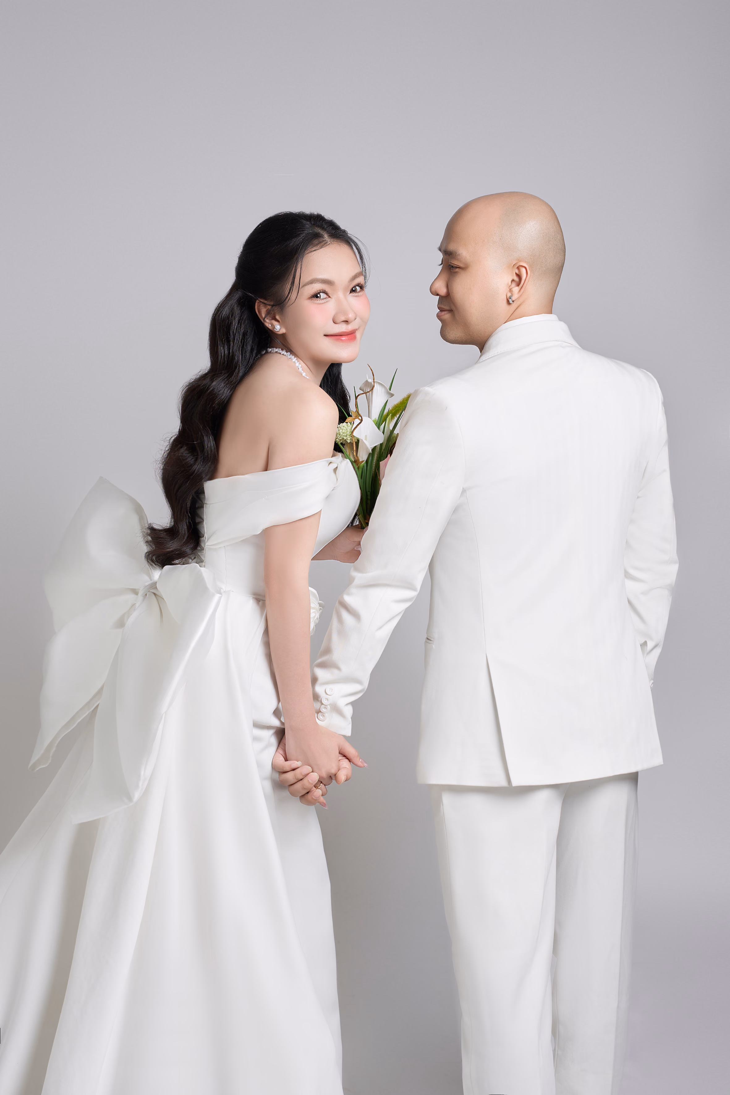 Nhìn từ phía sau, chú rể vest trắng nắm tay cô dâu váy trắng nơ lớn; cô dâu ngoái đầu lại mỉm cười, nền xám nhạt.