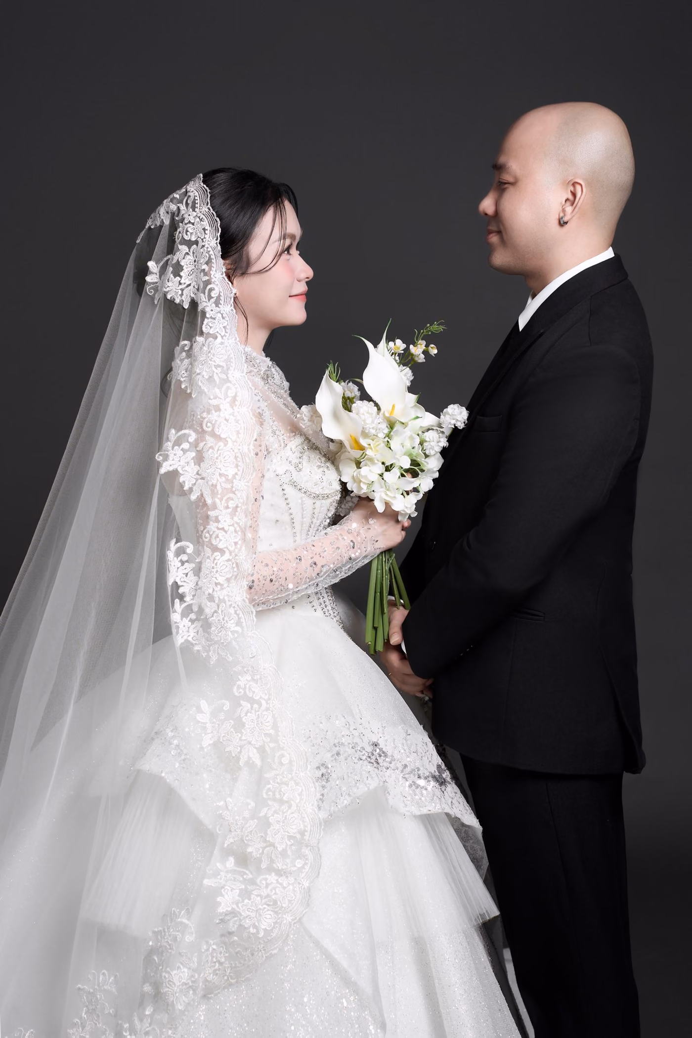 Cô dâu đội voan dài viền ren, váy cưới xếp tầng đính kim sa, cầm bó hoa calla trắng, đứng đối diện chú rể vest đen; hai người mỉm cười nhìn nhau, nền xám than.