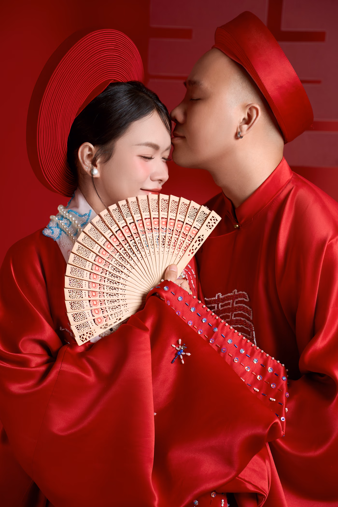 Cô dâu và chú rể trong áo dài đỏ và khăn đóng đỏ trước nền đỏ có chữ song hỷ; chú rể nhắm mắt hôn lên tóc cô dâu, cô dâu mỉm cười cầm chiếc quạt gỗ chạm hoa.